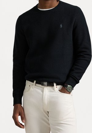 Mann trägt einen schwarzen Langarmpullover mit kleinem Logo, weiße Hose, schwarzen Gürtel und eine Armbanduhr mit grünem Zifferblatt.