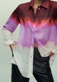 Persona che indossa una camicia a maniche lunghe ombré rossa, rosa, viola e bianca con i polsini arrotolati e pantaloni di pelle nera, mani in tasca.
