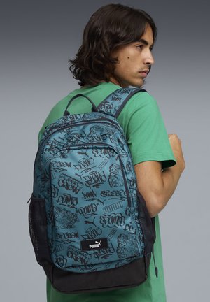 Joven con camisa verde que lleva una mochila azul de Puma con patrones en estilo grafiti negro sobre un fondo gris.