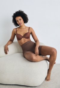 Καφέ σουτιέν με ρυθμιζόμενους ιμάντες και ελαφριά επένδυση, σε συνδυασμό με καφέ ψηλόμεσο shapewear. Μοντέλο καθισμένο σε έναν υφασμάτινο, ανοιχτόχρωμο καναπέ.