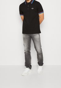 Zwarte poloshirt met een blauw en rood geribde kraag en manchetten, gecombineerd met vervaagde grijze jeans en witte sneakers. Eenvoudig ontwerp, moderne pasvorm.