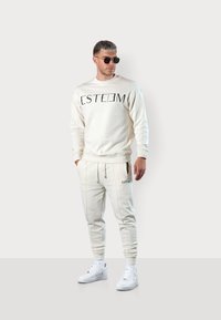 Beige sweatshirt with black "ESTEEM" text, paired with matching joggers. Features ribbed cuffs, side pockets, and white sneakers.