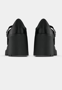 Μαύρα παπούτσια Mary Jane με χοντρό τακούνι και στρογγυλεμένα τακούνια, ανώτερο μέρος από λείο δέρμα και λουράκια με αγκράφα, εμφανίζονται από πίσω σε λευκό φόντο.