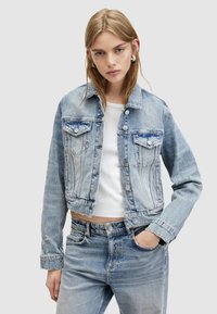 Giacca in denim azzurro chiaro, tagliata, con due tasche sul petto, bottoni metallici argento e dettagli sbiaditi. Abbinata a una camicia bianca e jeans.