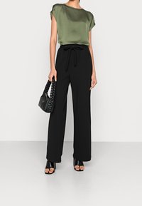 Grüne Satinbluse mit kurzen Ärmeln, kombiniert mit schwarzen, weit geschnittenen Hosen mit gebundener Taille. Accessoires sind eine geflochtene schwarze Handtasche.