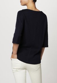 Suéter de punto trenzado azul marino de mangas tres cuartos, con un escote amplio y un patrón texturizado, llevado sobre jeans blancos.