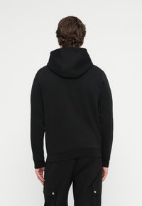 Mężczyzna z falującymi włosami widziany z tyłu, ubrany w czarną bluzę z kapturem i czarne spodnie cargo na jednolitym białym tle.