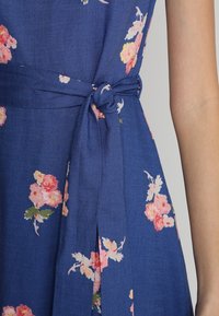 Blå blommig klänning med rosa och gula blommor, tillverkad av lättviktsmaterial. Knytning vid midjan som accentuerar med flytande kjoldetalj.