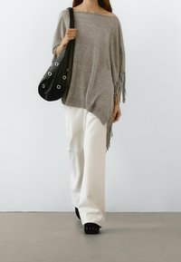 Grauer gestrickter Poncho mit Fransen, kombiniert mit weißen weit geschnittenen Hosen und einer schwarzen Tasche mit runden Ösen. Schwarze Slipper.