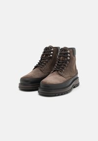 Stivaletti alla caviglia in suede marrone presentano una suola in gomma nera, occhielli in metallo e lacci. Un dettaglio in pelle liscia sul tallone arricchisce il design.