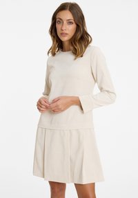 Beiges Rippstrickkleid mit langen Ärmeln; hat eine eng anliegende Oberseite und einen plissierten Rock, der Textur und klare Linien präsentiert.