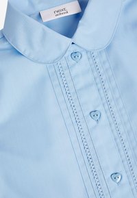 Helles blaues Schulhemd mit rundem Kragen, herzförmigen Knöpfen und dekorativen Nähten an der Vorderseite. Glattes, leichtes Material.
