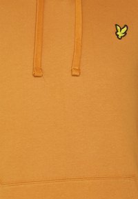 Haut à capuche orange en tissu doux, avec une poche kangourou, des cordons de serrage et un petit logo jaune brodé sur la poitrine.