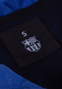 Blauwe stof met een gaasstructuur. Een zwart etiket toont de letter "S" en een grijs embleem met verticale strepen en een voetbal.