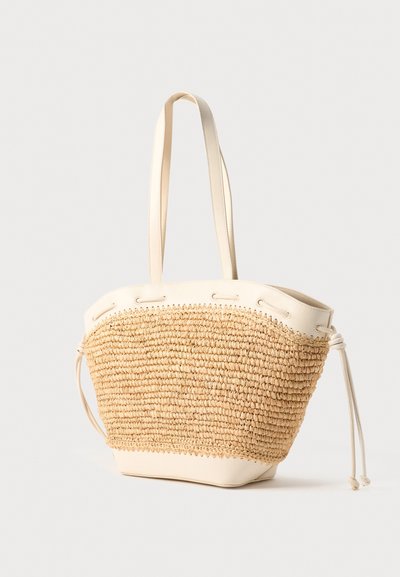 Πλεκτή τσάντα από άχυρο με κρέμ δερμάτινες λεπτομέρειες, πλευρικές κορδόνια και διπλούς ιμάντες ώμου σε ουδέτερο φόντο.