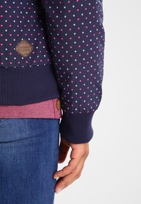 Manche bleu marine avec des pois roses et blancs, bord-côte, partie d'un pull rose, et main reposant près d'un jean bleu.