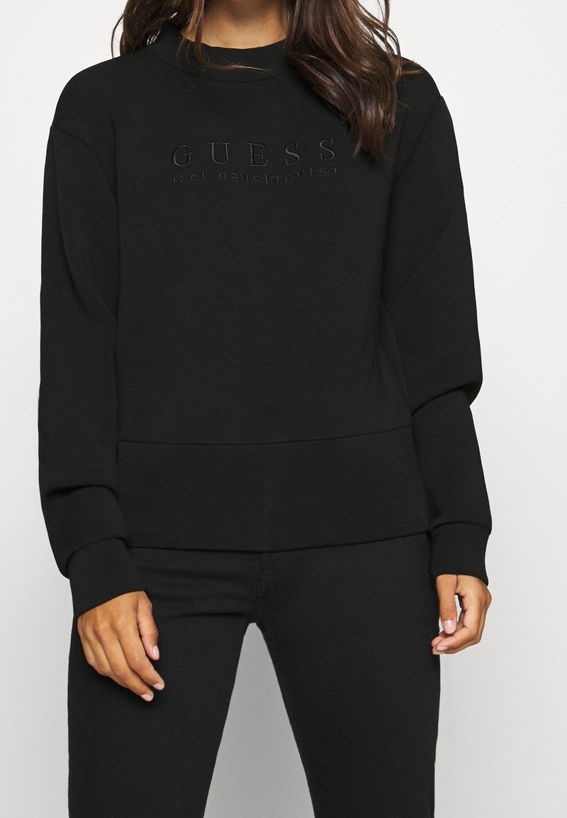 Femme portant un sweat-shirt noir avec le mot "GUESS" embossé sur la poitrine, associé à un pantalon noir, sur un fond uni.