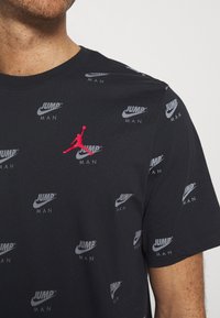 Camiseta de algodón negra con logotipos de Nike en gris repetidos y un destacado logotipo de Jumpman rojo en el pecho; cuello redondo y mangas cortas.