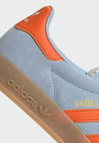 Light blue suede sneaker with orange accents, featuring three stripes and a transparent rubber sole. Text "Gazelle" and Adidas logo present.