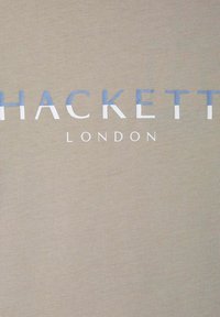 Bež pamučna majica s rebrastom teksturom. Ispisano "HACKETT" u plavoj i bijeloj boji, sa "LONDON" ispod u manjim bijelim slovima.