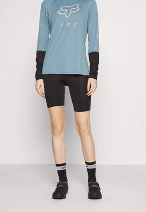 Personne portant un maillot de cyclisme Fox bleu à manches longues, un cuissard rembourré noir, des chaussettes noires et des chaussures de cyclisme noires, debout contre un mur uni.