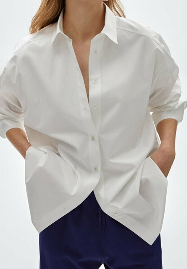 Chemise blanche boutonnée en tissu léger, présentant une coupe décontractée, des épaules tombantes et de grandes poches. Texture lisse et design net.