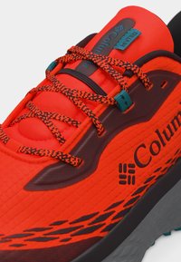 Φωτεινό πορτοκαλί παπούτσι για τρέξιμο σε μονοπάτια Columbia με κορδόνια σε πορτοκαλί και μαύρο μοτίβο, μαύρη σόλα και στήριγμα κορδονιών και ετικέτες λογότυπου σε τιρκουάζ.