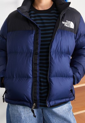 Personne portant une doudoune The North Face bleue et noire par-dessus une chemise noire à rayures bleues avec un jean bleu clair.