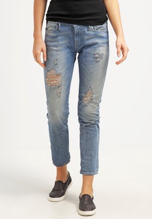 Kobieta ubrana w jasnoniebieskie, podarte jeansy, czarne buty wsuwane i czarną bluzkę, stojąca na prostym białym tle.