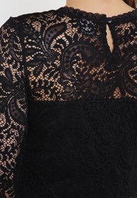 Top in pizzo nero con motivi floreali intricati, una parte superiore trasparente e una parte inferiore in tessuto solido con chiusura a bottone sul retro.
