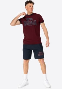 Burgundi medvilnės marškinėliai su tamsiu grafiniu logotipu, derinami su tamsiai mėlynomis šortais, turinčiais atitinkamą logotipą. Avėti su baltais sportbačiais ir kojinėmis.