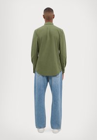 Olivgrön långärmad skjorta med en veck bak, rundad nederkant, knäppningar vid ärmsluten, kombinerad med ljusblå vida jeans och vita sneakers.