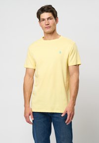 Tricou din bumbac galben cu mânecă scurtă, având un logo teal pe pieptul stâng, guler rotund și o croială lejeră. Blugii sunt de un albastru închis.