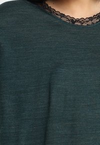 Dunkelgrüner Strickoberteil mit Rundhalsausschnitt, der mit schwarzer Spitzenborte verziert ist und den Kontrast zwischen Texturen und Farben betont.