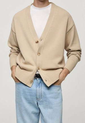Cárdigan beige abotonado usado sobre una camiseta blanca, combinado con vaqueros azul claro, manos metidas en los bolsillos, solo se muestra el torso.