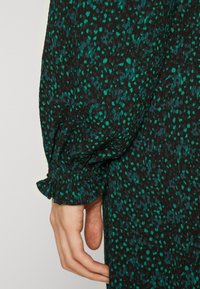 Svart tyg med grönt stänkmönster; har en veckad, rynkad mudd och texturerad yta. Närbild på en hand som håller tyget.