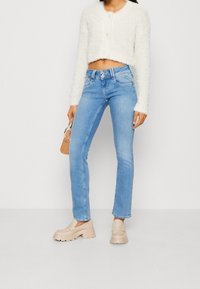 Žena v světle modrých dvojoboučkových džínách, krémovém chlupatém krátkém svetru, béžových masivních mokasínech, držící malou hnědou kabelku proti prostému pozadí.