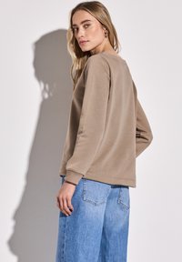 Světle hnědá mikina s dlouhými rukávy, kulatým výstřihem a manžetami, zkombinovaná s volnými modrými džíny. Hladká textura materiálu.