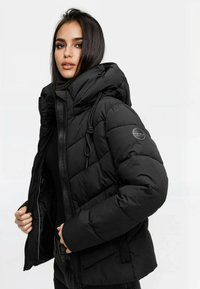 Mujer joven vestida con una chaqueta de plumas negra acolchada con capucha, sosteniendo la parte delantera sin zip abierta, de pie contra un fondo blanco liso.