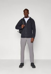 Črna jakna iz flisa z zadrgo, sive hlače in črni pohodniški čevlji. Model nosi belo majico pod jakno in ima črn nahrbtnik.