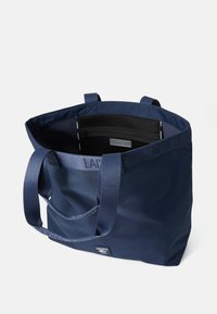 Marineblå tote-veske med kraftig stoff, doble håndtak og en svart glidelås lomme inni. Har preget logo-merke på utsiden.
