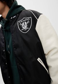 Zwarte en roomkleurige varsityjack met een Raiders-logo, met een soepele textuur, drukknoopsluitingen en gestreepte zakaccenten.