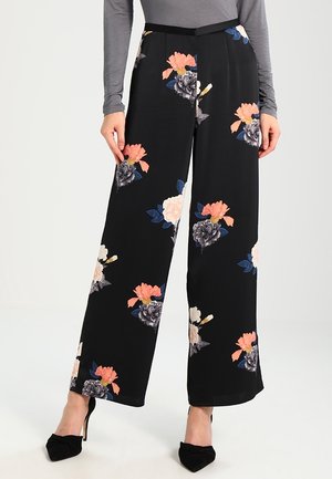 Zwarte wijde-pantalon met grote roze, witte en grijze bloemmotieven, gedragen met een grijze lange mouwen top en zwarte puntige hakken.