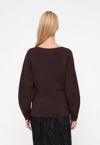 Maglione oversized marrone scuro con scollatura ampia e maniche lunghe, abbinato a una gonna nera texturizzata con un motivo a foglia.