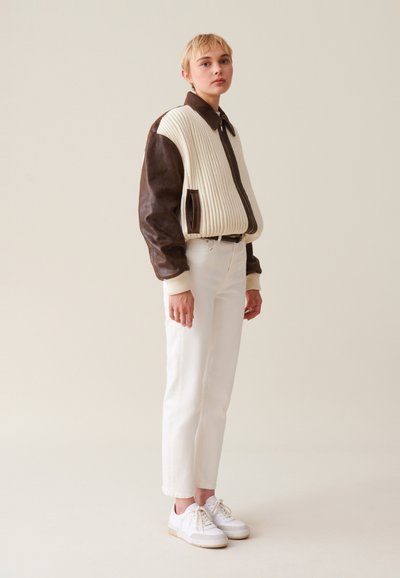Hnedá kožená bunda s krémovým ribovaným materiálom a golierom, v kombinácii s bielymi nohavicami a bielymi teniskami. Jednoduchý, štíhly dizajn.