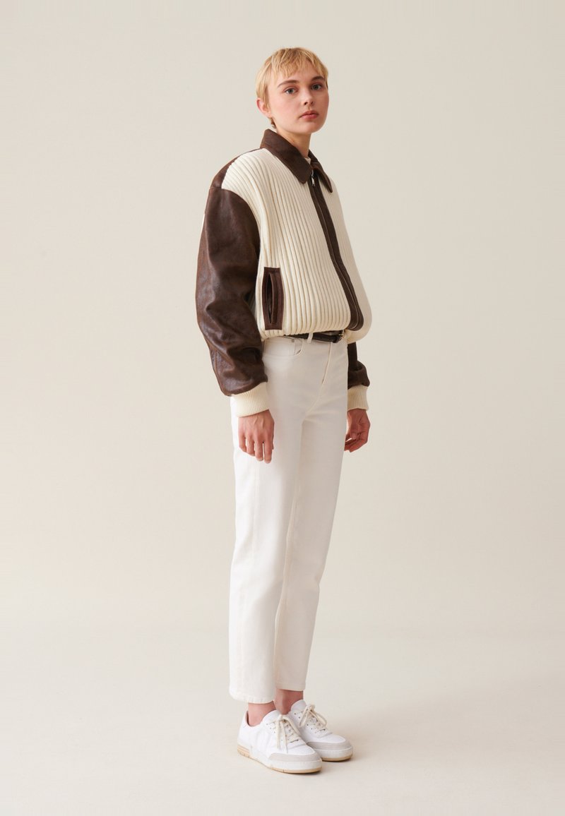 Braune Lederjacke mit cremefarbener, gerippter Stoffeinsatz und Kragen, kombiniert mit weißen Hosen und weißen Sneakern. Einfaches, klar strukturiertes Design.