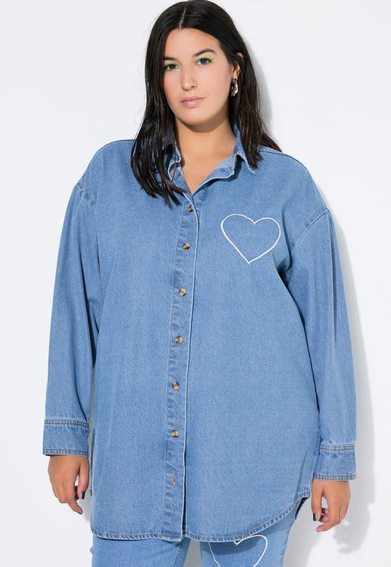 Lichtblauw denimshirt met lange mouwen, knoopsluiting aan de voorkant en een hartvormige witte borduursel op de linkerborst. Voorzien van een klassieke kraag.