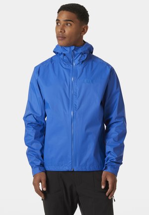 Man draagt een felblauwe gewatteerde jas met capuchon en ritssluiting, staand voor een effen lichte achtergrond, kijkt lichtjes opzij.