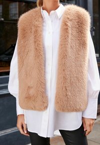 Gilet en fausse fourrure beige avec une texture douce, de larges rabats et sans fermeture, porté sur une chemise blanche à boutons et un legging noir.