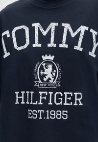 Ναυτικό ύφασμα με φθαρμένο λευκό κείμενο "Tommy Hilfiger Est. 1985" και κεντρική απεικόνιση με λιοντάρι και τις λέξεις Palm Springs, California, New York.
